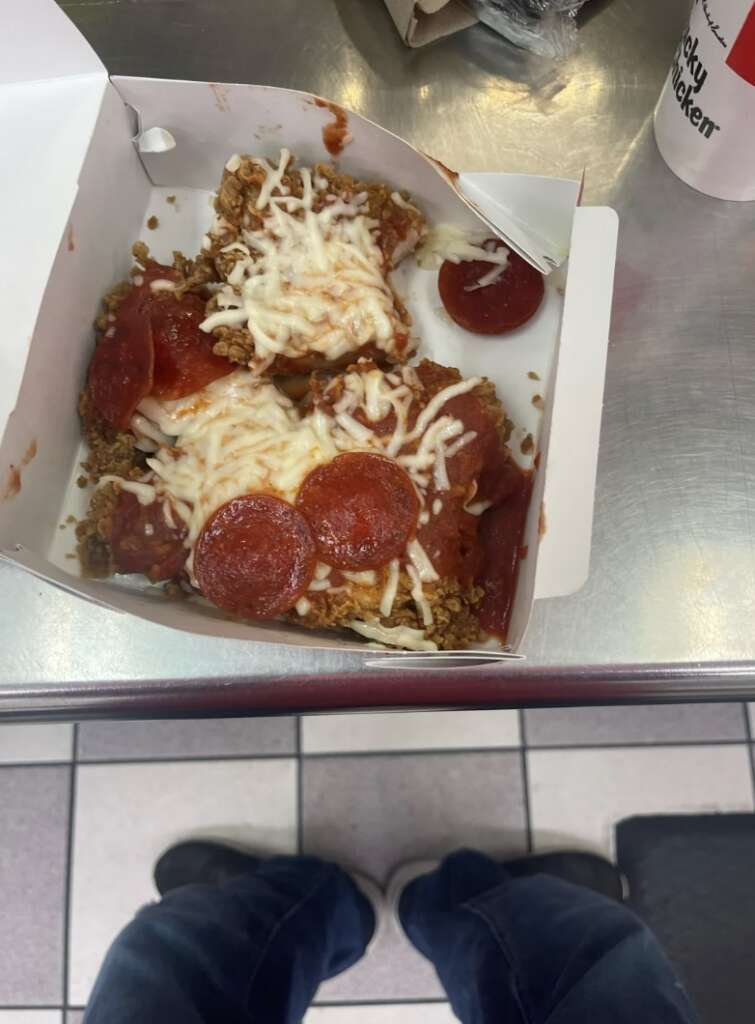 A to-go box containing a dish of fried chicken topped with melted cheese, tomato sauce, and pepperoni slices. The box rests on a stainless steel table. Nearby on the table is an unopened plastic cutlery pack and a drink cup.