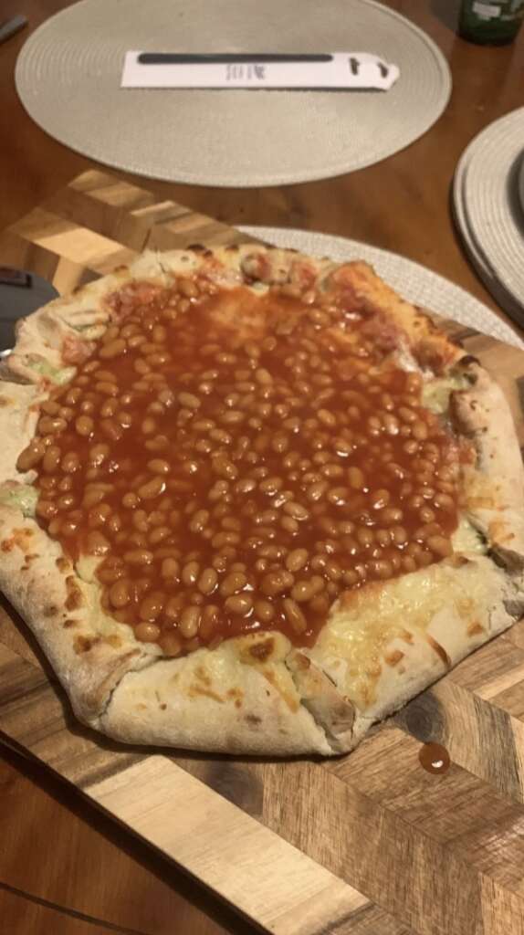 A pizza topped with baked beans on a wooden board. The crust is lightly cooked, and the beans cover the melted cheese underneath. A pizza cutter is partially visible on the left. The setup is on a wooden table with placemats.