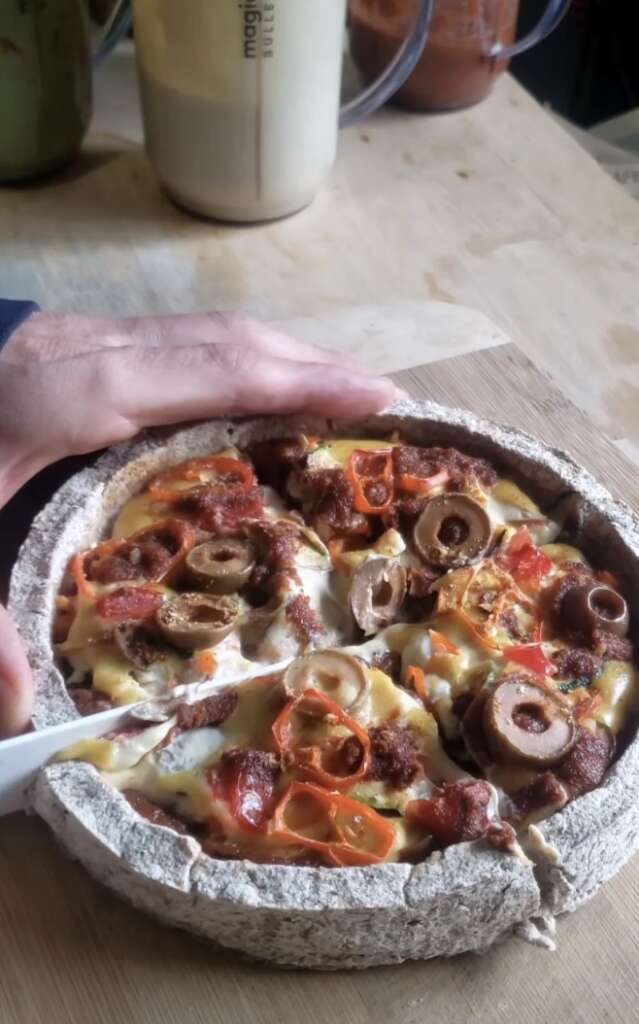 A person is cutting a pizza with a knife. The pizza is topped with olives, cheese, sliced bell peppers, and chunks of meat. It is on a round wooden board, and a hand is visible on the left.