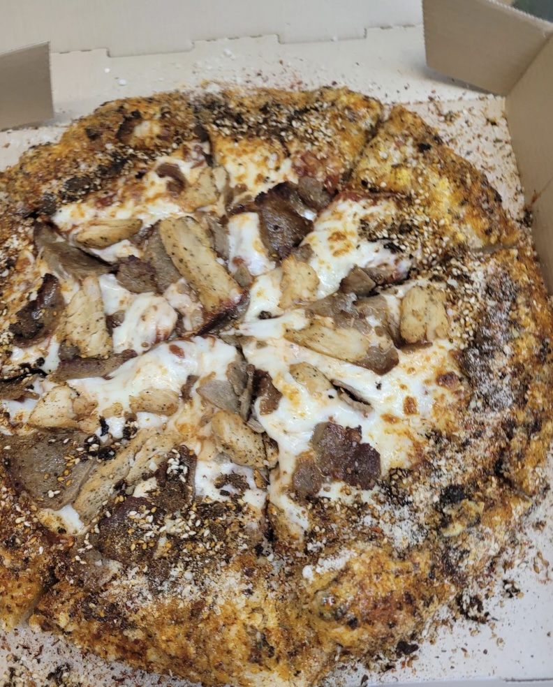 A pizza with a thick crust topped with slices of seasoned meat and melted cheese, sprinkled with herbs and spices. It is cut into slices and displayed inside a cardboard box.