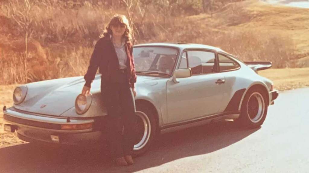 A person stands beside one of the coolest cars from the 1980s—a vintage silver sports car with wide rear fenders and a rear spoiler. It's parked on a paved road, surrounded by dry grass and hills, all bathed in warm sunlight.