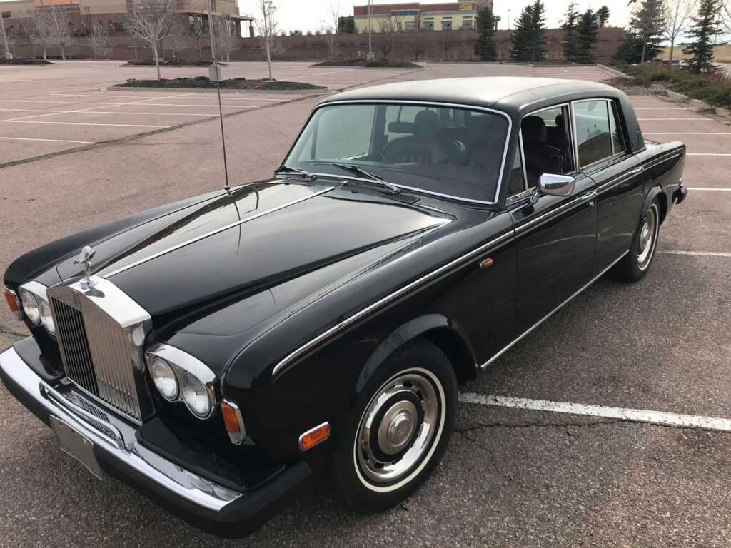 A classic black Rolls-Royce sedan, one of the coolest cars from the 1980s, is parked in an empty lot. Its shiny exterior and vintage design are highlighted with chrome accents and a distinct grille. The sky is overcast, and trees are visible in the background.