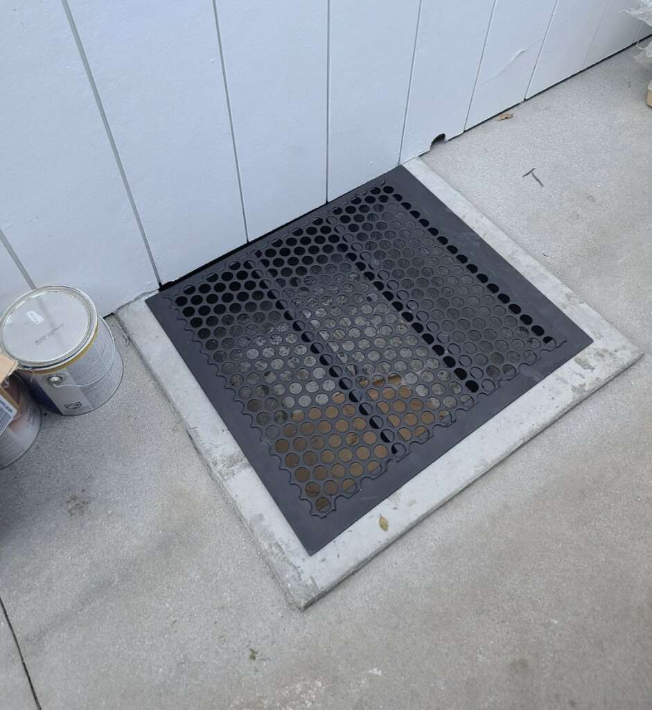 A black metal grate covers a ground opening next to a white wall, potentially part of recent structural assessments. Two paint cans sit nearby on the concrete surface.