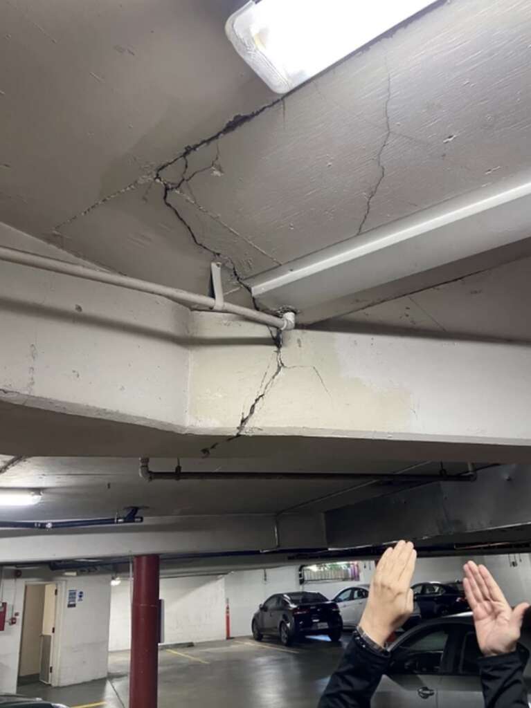 A cracked concrete beam in a parking garage is carefully examined during structural assessments, with two hands measuring the gap. Nearby, several cars are parked as a fluorescent light casts an eerie glow from the ceiling, highlighting the beam's significant damage.