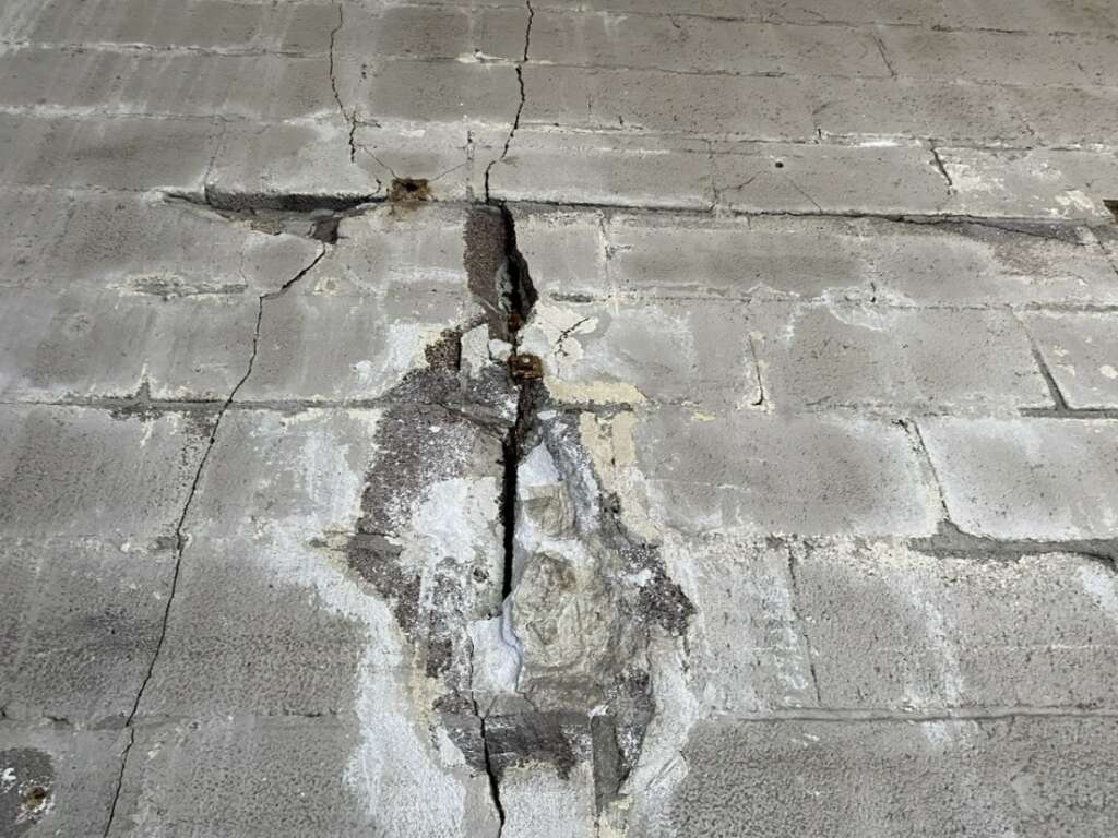 A cracked concrete wall with visible damage reveals deep fractures and areas of erosion, highlighting the need for thorough structural assessments. The weathered surface displays uneven textures and varying shades of gray.