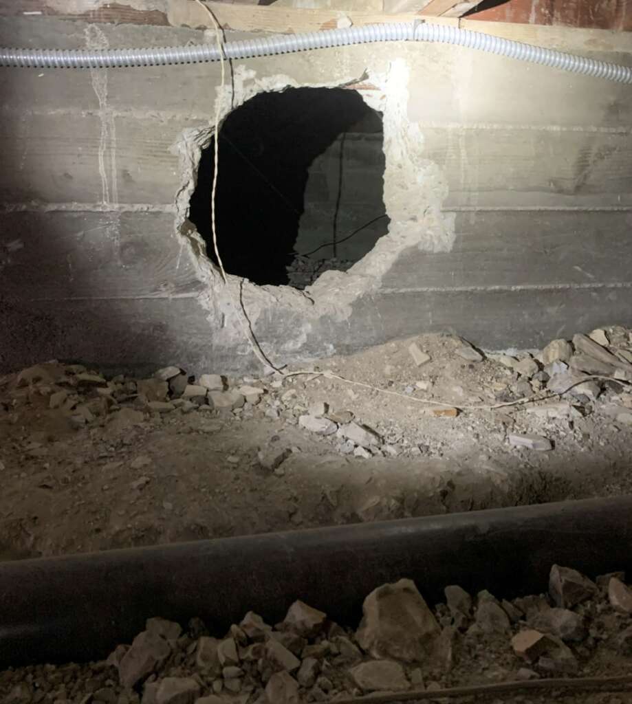 A large hole in a concrete wall, surrounded by rubble and debris, is illuminated by a flashlight. Nearby, a plastic pipe and some wires are visible. This scene resembles the aftermath of structural assessments in a dimly lit basement or crawl space.