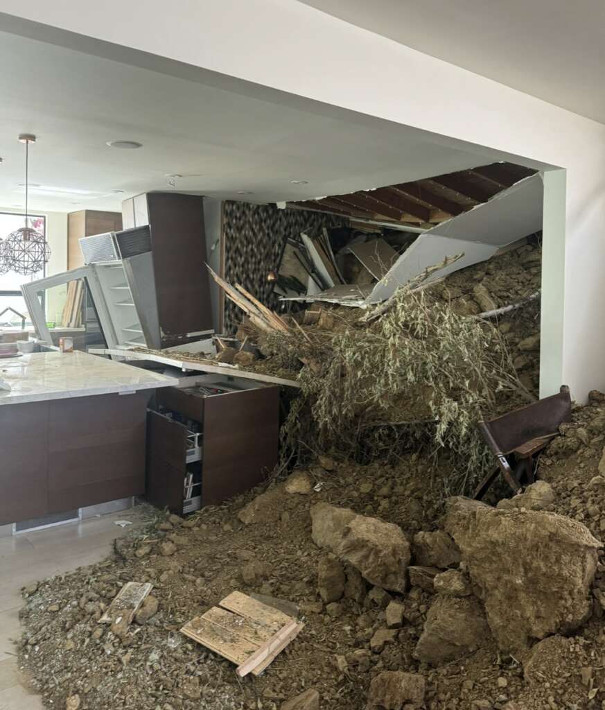 The image shows a kitchen heavily damaged by a landslide, highlighting the need for structural assessments. Dirt, rocks, and debris fill the area, with overturned furniture and appliances. A tree branch is visible among the rubble. The ceiling and walls are also affected by the damage.