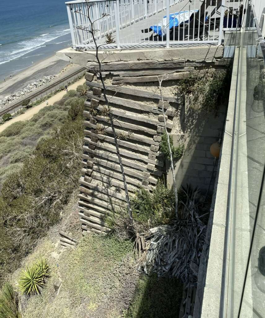 A view of a seaside cliff with a wooden retaining structure supports a coastal walkway, carefully maintained through regular structural assessments. The ocean and beach are visible in the background, alongside some plants and a glass fence.