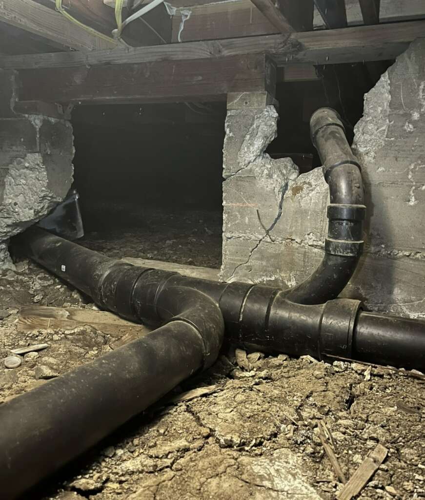 A basement with an exposed dirt floor and two large black pipes running through it. The walls, made of concrete, show signs of damage and cracks. Dim lighting highlights the rough texture of the surroundings, warranting a structural assessment to ensure stability and safety.