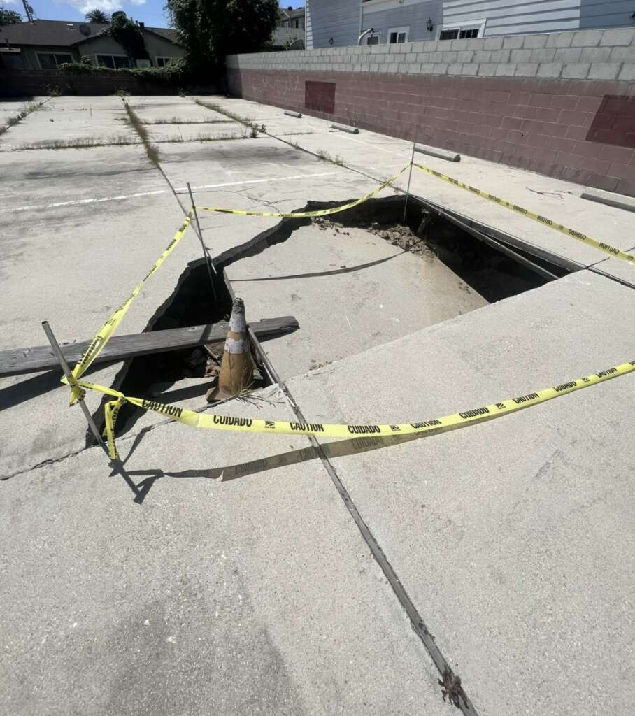 A large hole in the concrete surface, possibly the focus of ongoing structural assessments, is surrounded by caution tape. Wooden beams frame the area, and a traffic cone sits within. Nearby, a brick wall and some houses provide an urban backdrop to this scene of inspection.