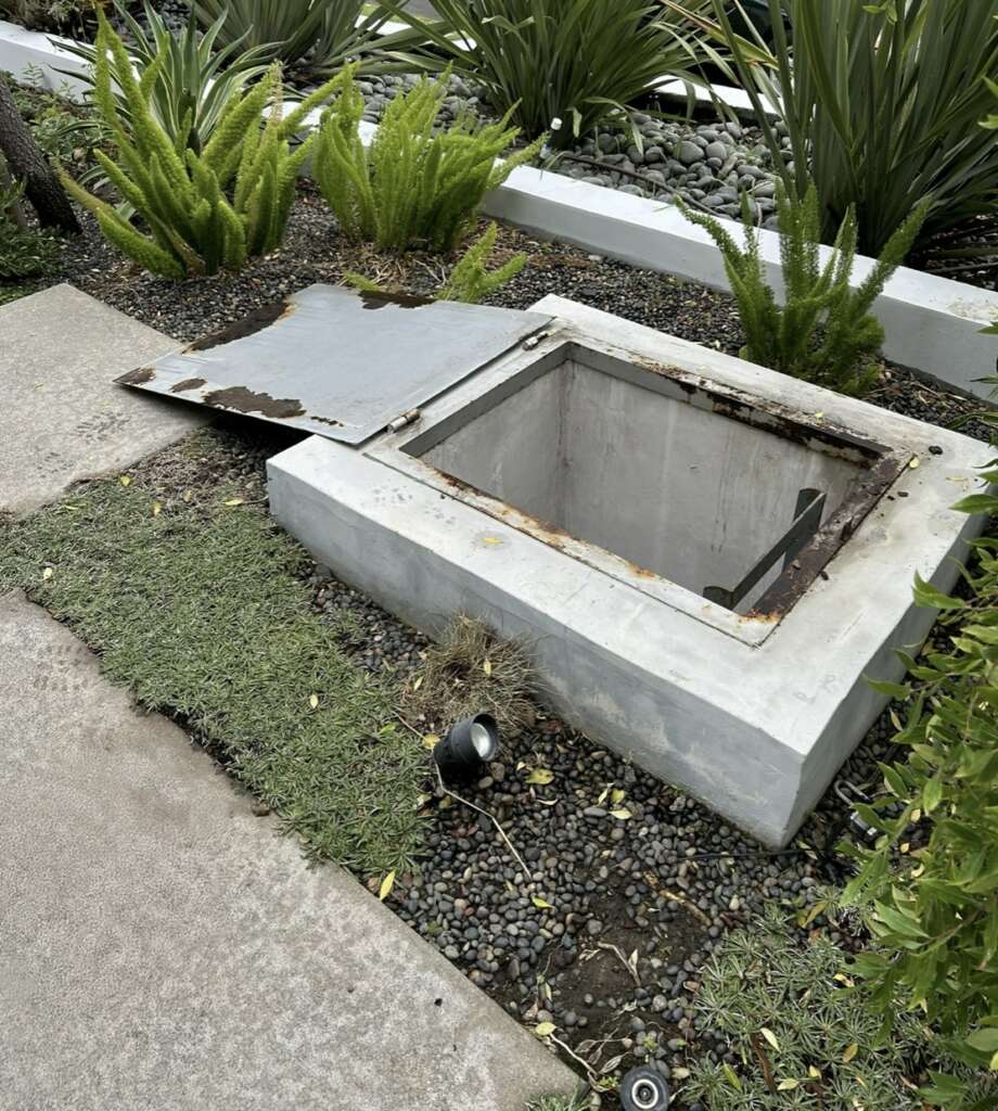 An open metal hatch on a concrete box, often subject to structural assessments, is surrounded by greenery and small decorative stones in a landscaped garden area. The metal cover is partially laid on the ground nearby.