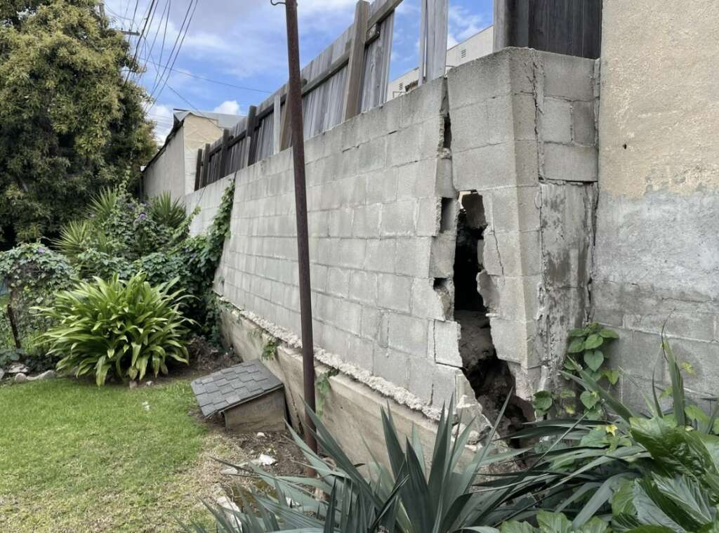 A cracked concrete wall with a large hole, pending structural assessments, is supported by wooden panels. Surrounding the wall are green plants and grass beneath a partly cloudy sky.