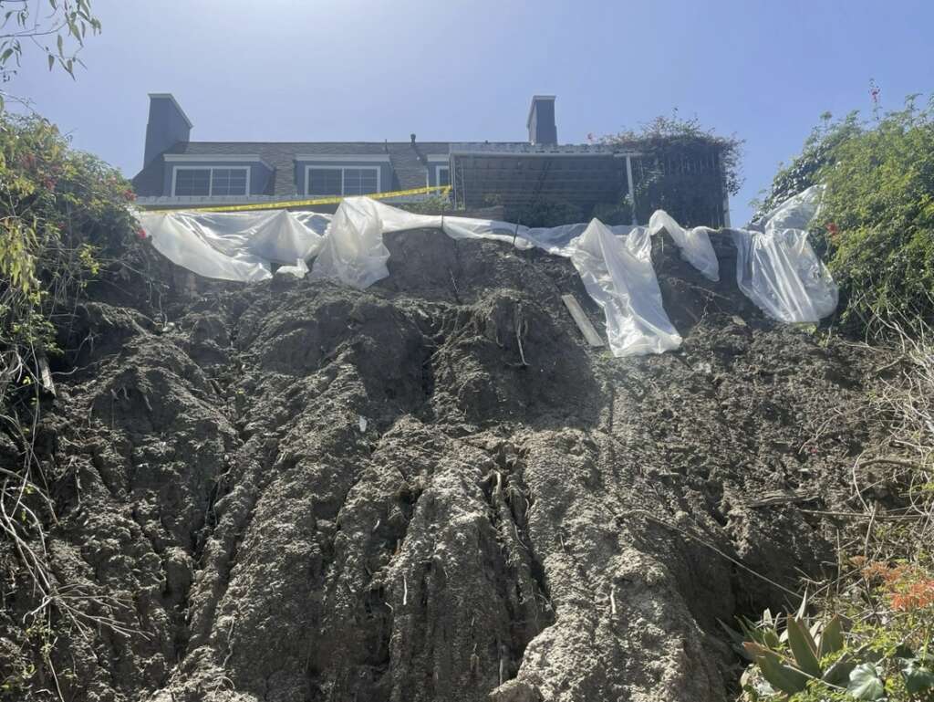 A house is precariously perched on the edge of a slope with a landslide beneath it, awaiting structural assessments. The top of the slope is covered with plastic sheets and caution tape, while vegetation flanks both sides under a clear sky.