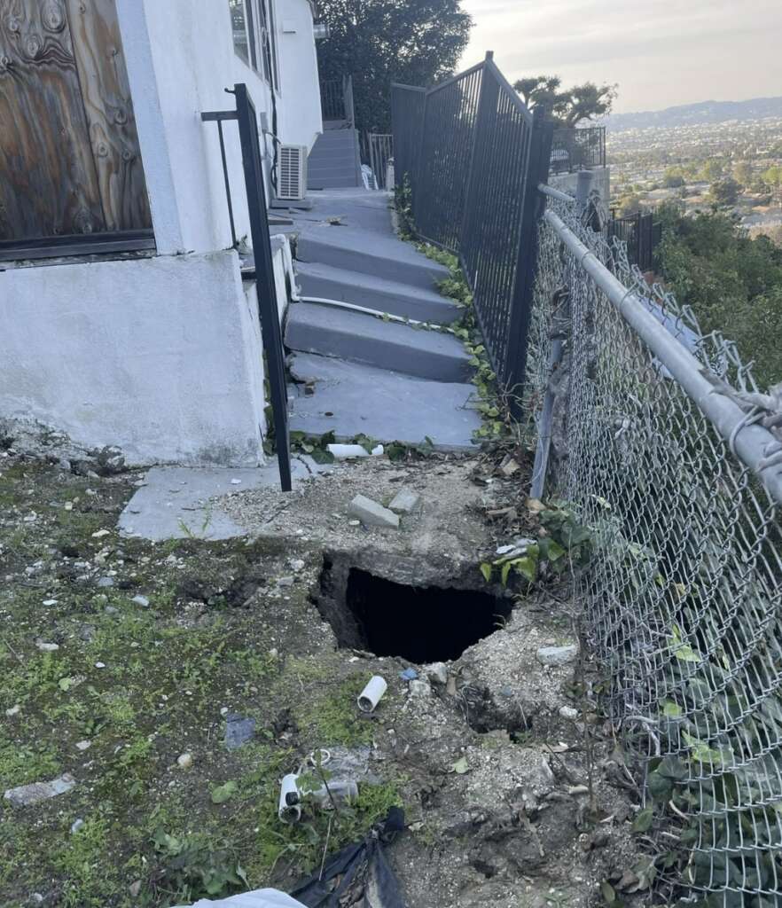 A hillside path with cracked grey stairs leads to a chain-link fence. Near the stairs, a large hole hints at the need for structural assessments, with scattered debris around it. The background features a distant city view under a clear sky.