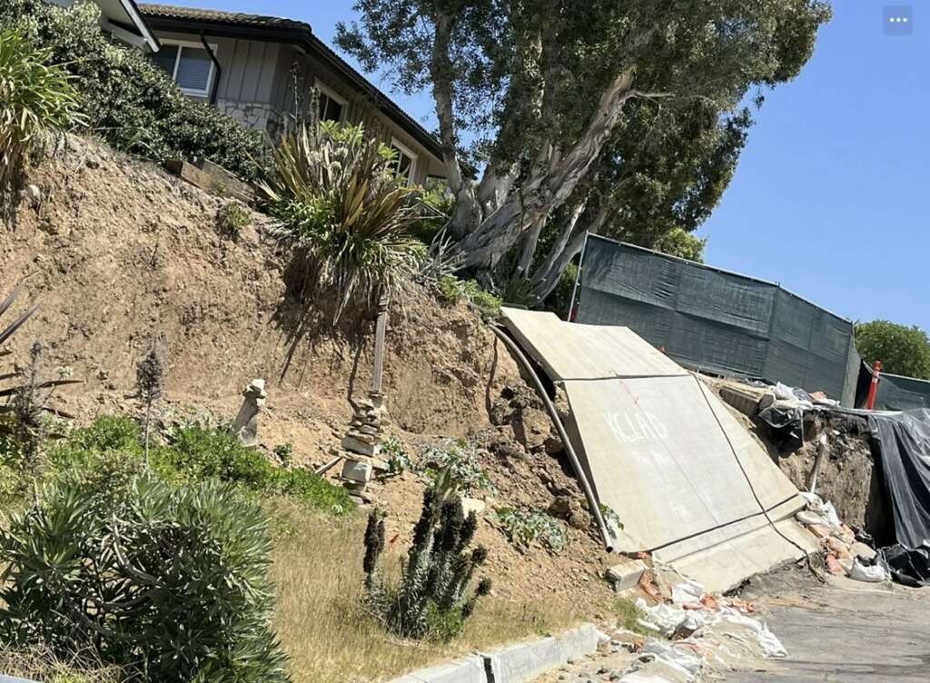 A house on a hilltop with visible erosion and a partially collapsed hillside undergoes structural assessments. Sandbags and barriers provide support, while vegetation and trees surround the area, with construction fencing visible in the background.