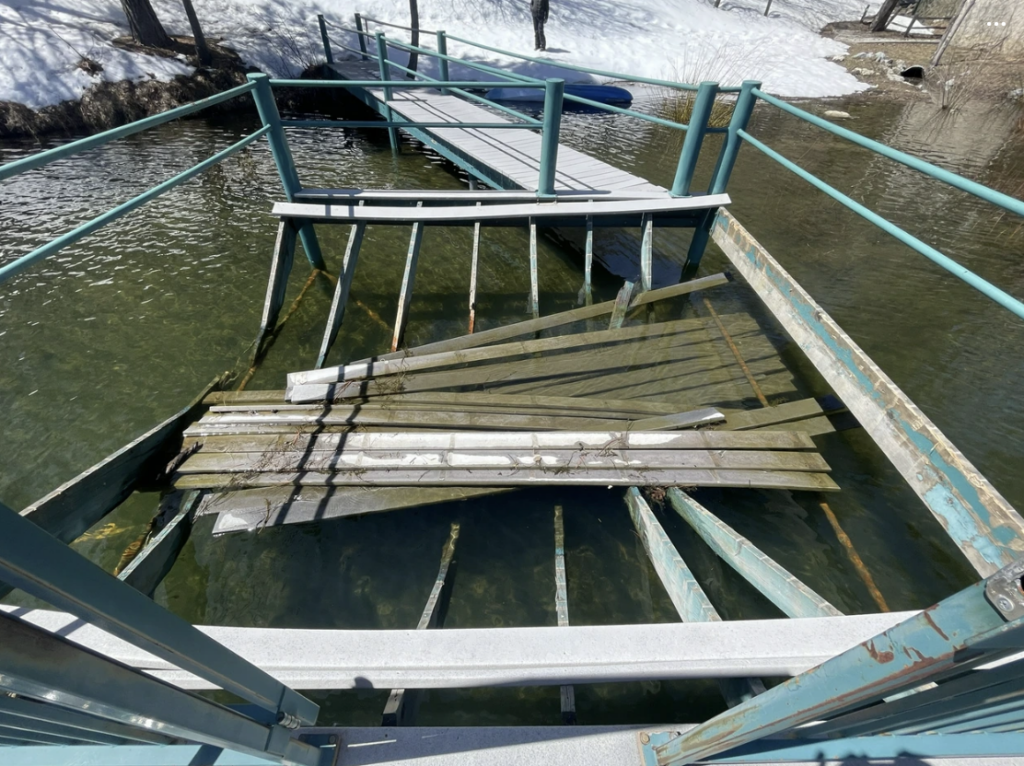 A broken footbridge with collapsed wooden planks spans a partially frozen body of water, beckoning the need for structural assessments. Snow blankets the surrounding ground, while metal railings frame the precarious bridge.