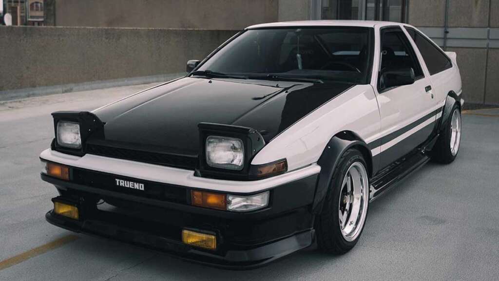 A sleek, vintage white and black sports car from the coolest cars of the 1980s era, with pop-up headlights and a spoiler, is parked in an outdoor setting. The design boasts distinctive angular lines and proudly displays "Trueno" on the front bumper.