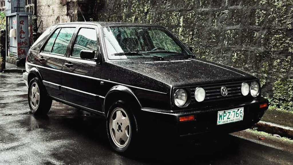 A vintage black Volkswagen hatchback, reminiscent of the coolest cars from the 1980s, is parked on a wet street. Its circular headlights and license plate reading "WPZ-766" gleam softly. The weathered stone wall in the background enhances the nostalgic atmosphere.