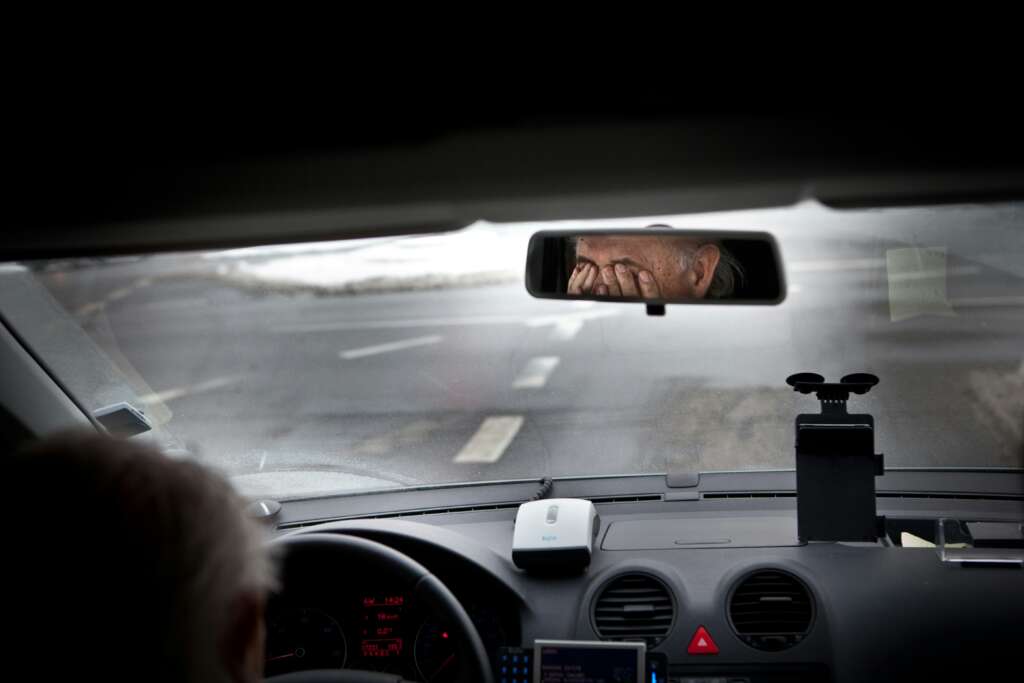 A person is driving a car, and their tired eyes are visible in the rearview mirror, reflecting exhaustion. The road ahead is empty, with a snowy landscape. The interior includes a dashboard with devices and a steering wheel.