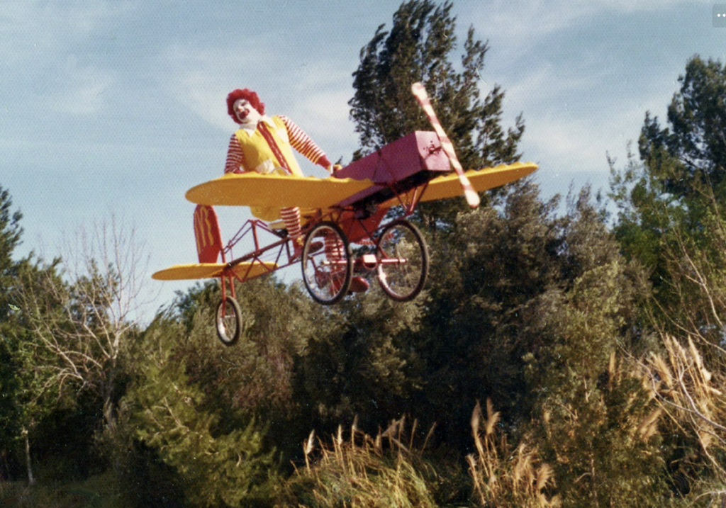 A person in a clown costume, with red and yellow stripes, flies a whimsical small yellow and red airplane-like contraption over a landscape with trees and grass, under a blue sky.