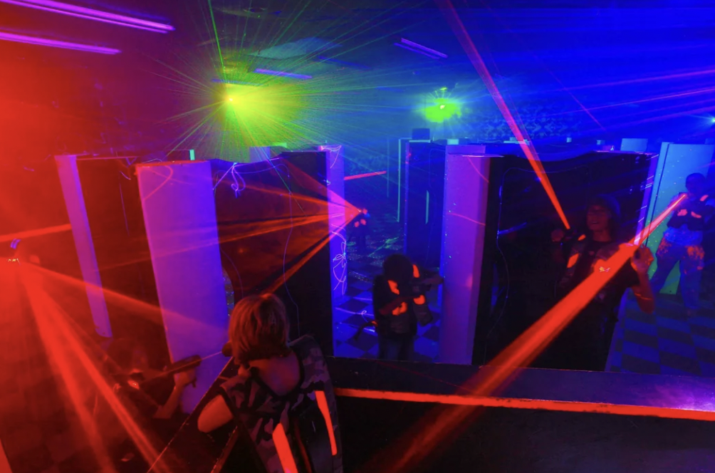 People playing laser tag in a dark room with vibrant red, green, and blue lights. Participants are wearing vests and aiming laser guns, surrounded by reflective surfaces and neon-colored lighting effects.