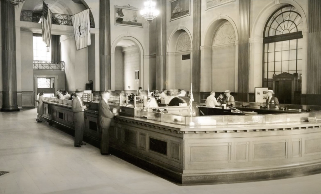 Vintage black-and-white photo of a busy bank interior. People are conducting transactions at a long wooden counter. The room features high ceilings, arched windows, and ornate columns. Flags hang from above, adding to the historic ambiance.