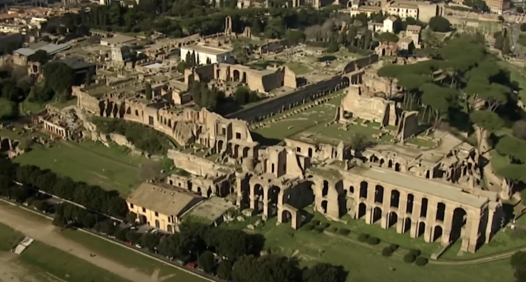 Aerial view of ancient Roman ruins, including large stone structures and arches surrounded by grassy areas. In the distance, modern buildings and trees are visible, contrasting with the historic architecture.