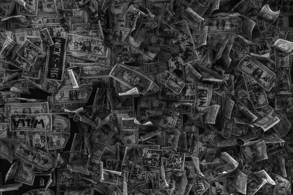 A chaotic pile of one-dollar bills, each marked with various handwritten messages and doodles, creating an overlapping collage. The image is in black and white, highlighting the texture and details of the currency and inked writings.