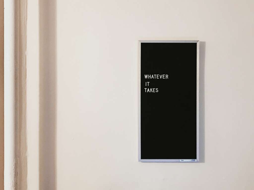 A black rectangular sign on a white wall displays the words "WHATEVER IT TAKES" in white capital letters.