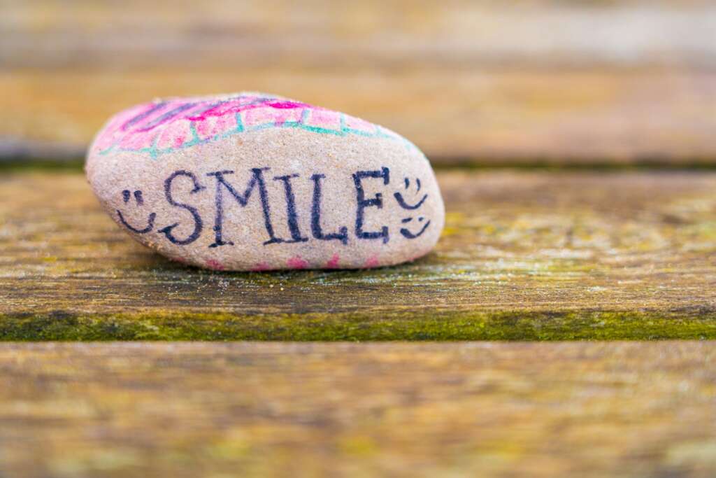 A smooth pebble with the word "SMILE" painted on it sits on a wooden surface. The lettering is in bold, dark blue, and is decorated with pink and green accents. The background is a textured wooden table.