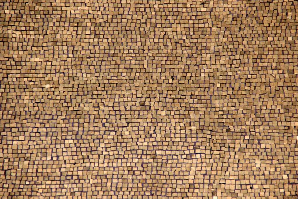 A close-up of a mosaic pattern made of small, square, tan-colored tiles. The tiles are unevenly placed, creating a textured and irregular surface across the image.