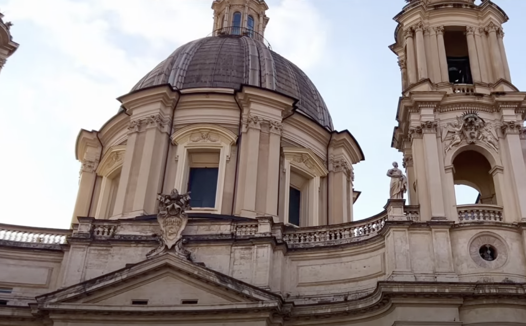 A close-up view of the ornate domes and architectural details of the Church of Sant'Agnese in Agone, located in Rome. The image shows the intricate carvings and classical design of the Baroque-style church against a clear sky.