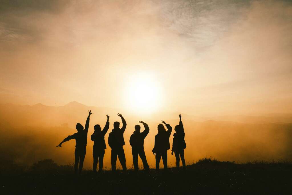 Silhouettes of seven people stand in a line with arms raised, facing a bright sunrise or sunset. They are on a grassy hill under a dramatic, cloudy sky, creating a sense of unity and celebration.