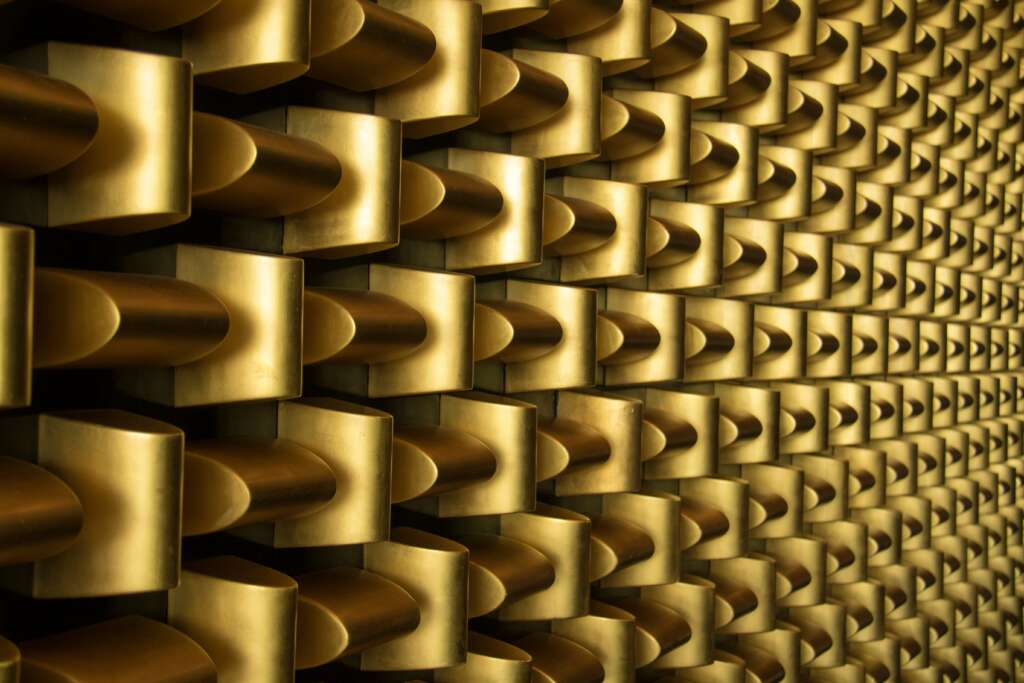 Close-up of an abstract gold-toned wall composed of a repeating 3D pattern of rectangular blocks, each featuring a protruding cylindrical knob in the center, creating a textured and symmetrical design.