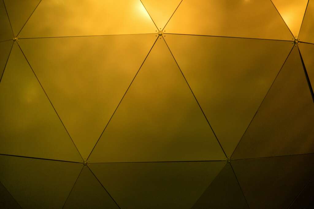 Geometric pattern of interconnected triangular panels under soft yellow lighting, creating a dome-like effect. The panels are reflective, with visible joints at each intersection.