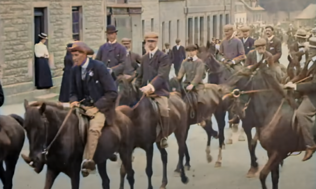 A historic scene shows a group of men and boys riding horses down a street, dressed in early 20th-century clothing, including hats and suits. The stone buildings and people on the sidewalk watching suggest a processional or parade event.