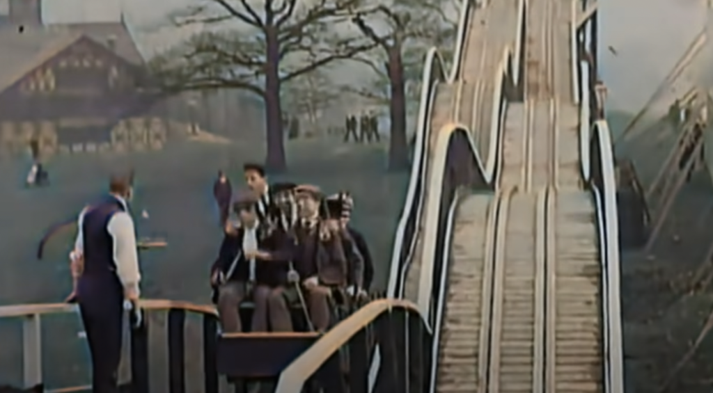 A vintage scene shows a steep wooden slide with people riding down in a cart. Two men in period attire with hats sit in the cart, ready to descend. A man stands nearby. Trees and a building are visible in the background.