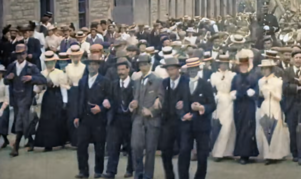 A vintage photo of a large crowd of people, mostly men and women in early 20th-century attire, walking arm-in-arm. Many are wearing hats and suits or long dresses. They appear to be participating in a peaceful demonstration or parade on a street.