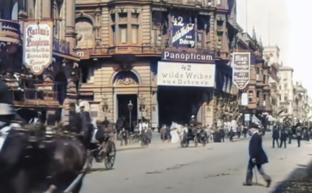 A bustling city street scene from the early 20th century with people walking, horse-drawn carriages, and a busy intersection. Buildings are adorned with signs, including "Panopticum" and "wilde Weiber." Architecture features ornate brickwork.