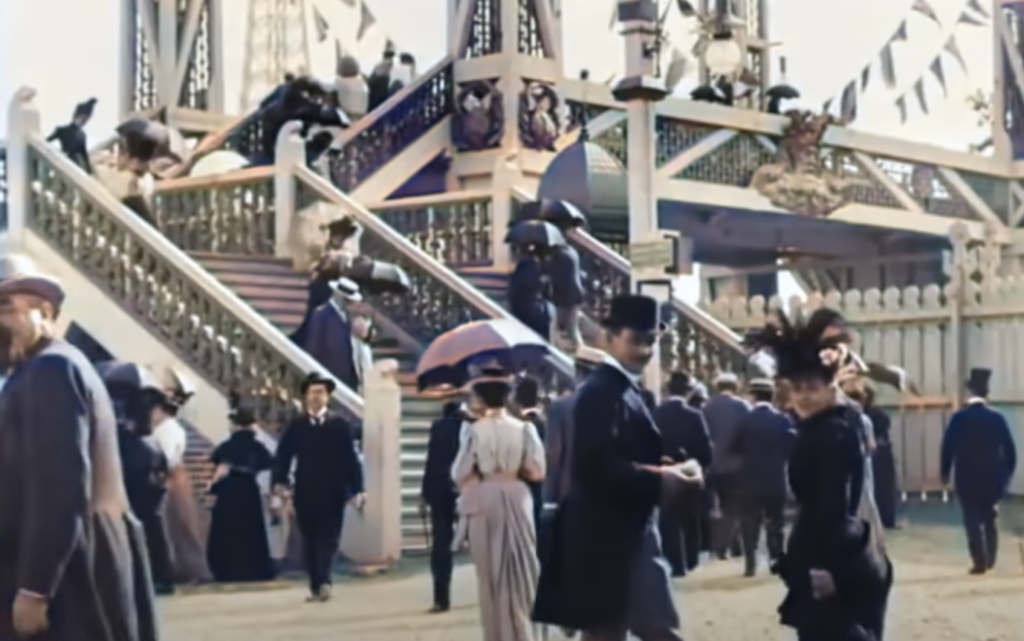 People dressed in late 19th or early 20th-century attire are gathered at an outdoor event. They are wearing hats and long coats, with women holding parasols. The scene includes ornate stairs and decorative flags.