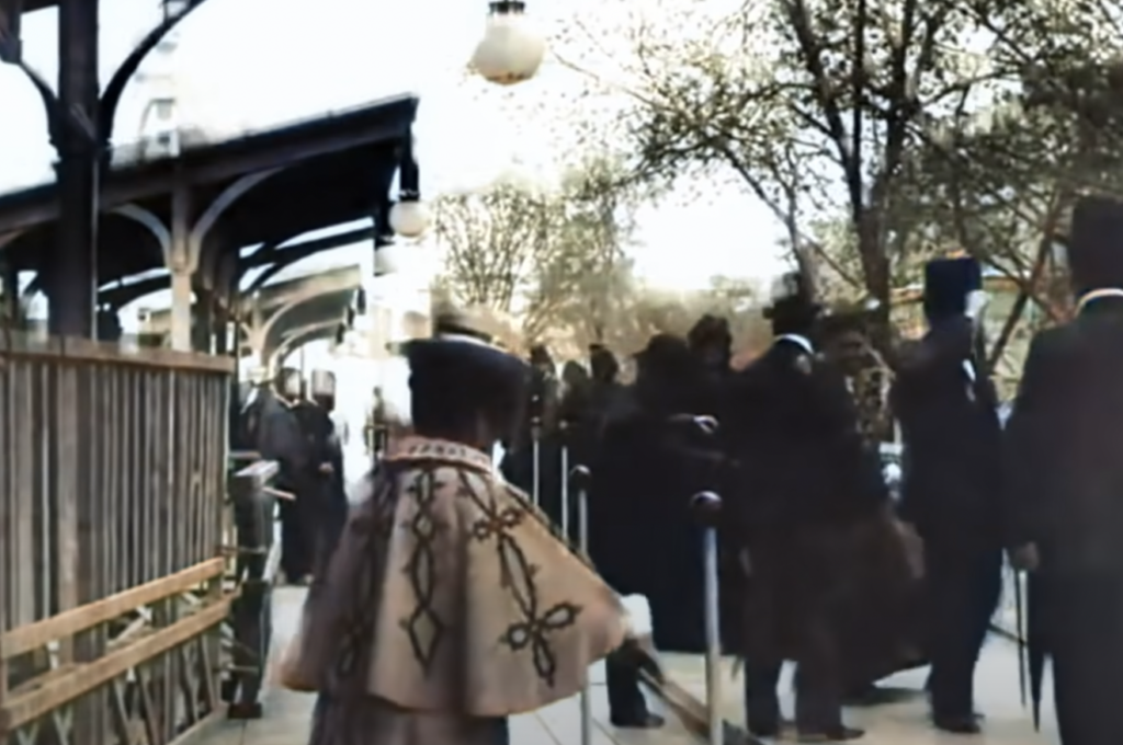 A scene at an early 20th-century train station shows people wearing period clothing, including top hats and capes, walking along a platform. Leafy trees line the background, and vintage lamps hang above the crowd.