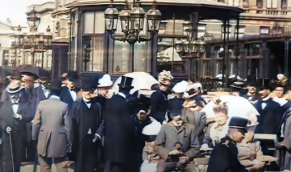 A bustling street scene from the early 1900s showing people in formal attire with hats and parasols outside a building with ornate street lamps. Men and women are engaged in conversation, capturing a moment of lively urban life.