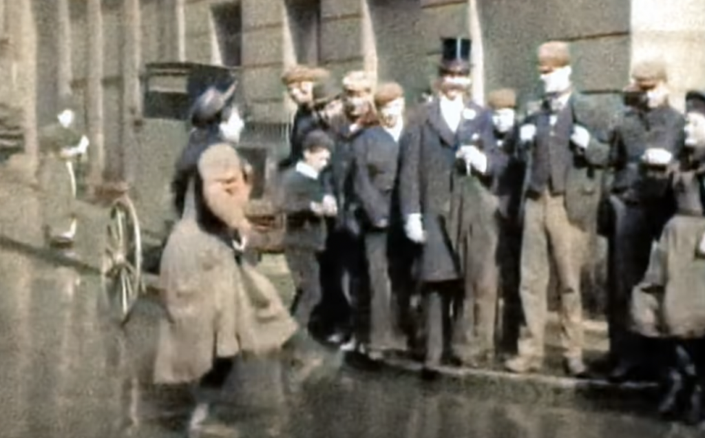 A historical street scene with a group of men and boys in early 20th-century attire, including hats and suits, gathered on a sidewalk. One man wears a top hat. A horse-drawn carriage and wet street are visible in the background.