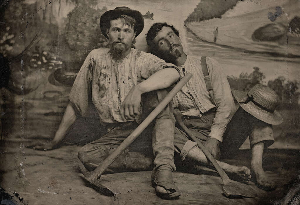 Two men with shovels sit on the ground in front of a painted backdrop depicting nature. Both wear hats and casual work clothes. One man leans on the other, suggesting camaraderie. The image has an old sepia tone.