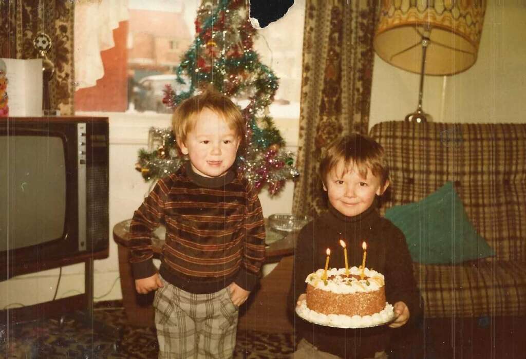 Two young children stand indoors in front of a decorated Christmas tree. One child is holding a cake with lit candles while the other stands beside him. A vintage TV and a lamp are in the background. Both kids are wearing sweaters.