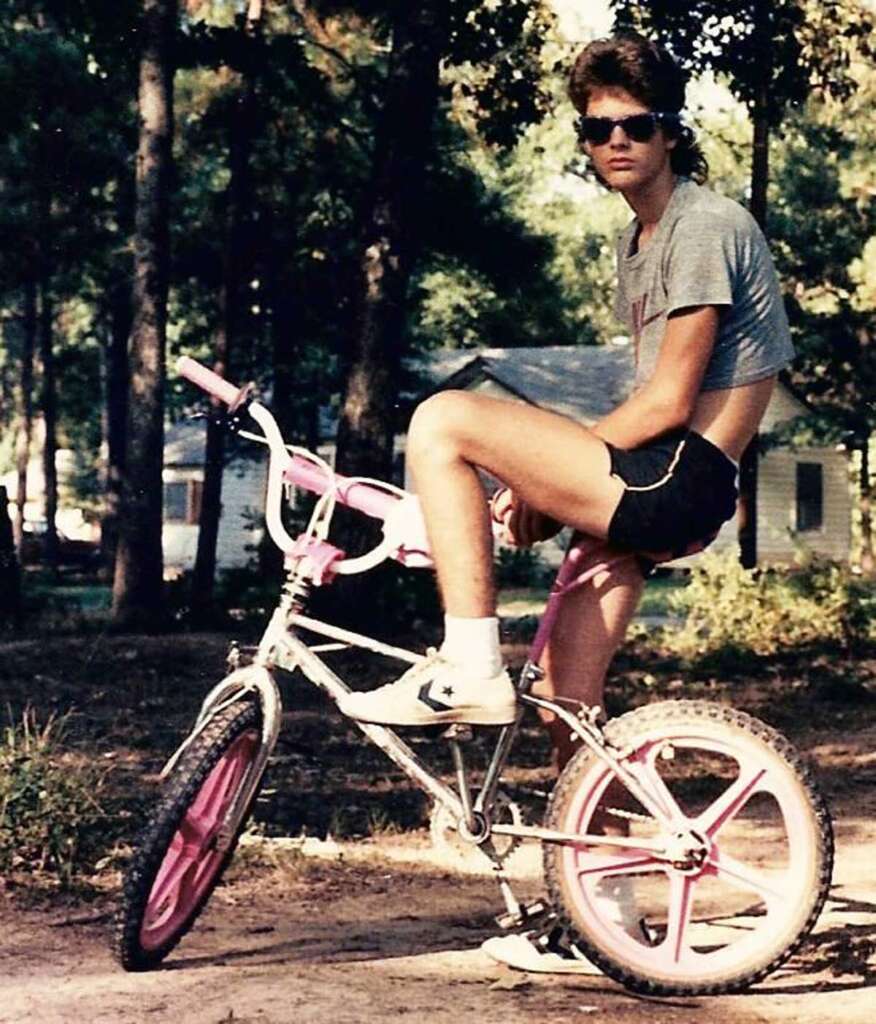 A person wearing sunglasses, a cropped grey t-shirt, and black shorts sits on a pink and white BMX bike. They are in a wooded area with trees and a small building in the background.