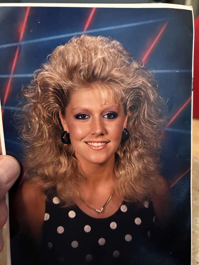 Portrait of a woman with voluminous blonde hair and bangs, wearing a black polka dot dress and hoop earrings. The backdrop features a retro-style neon light pattern with diagonal red and blue lines.