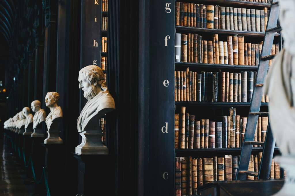 A grand library with tall, dark wooden shelves filled with old books. Busts of historical figures line the aisle, and a ladder leans against one of the shelves, suggesting a classic, scholarly atmosphere.