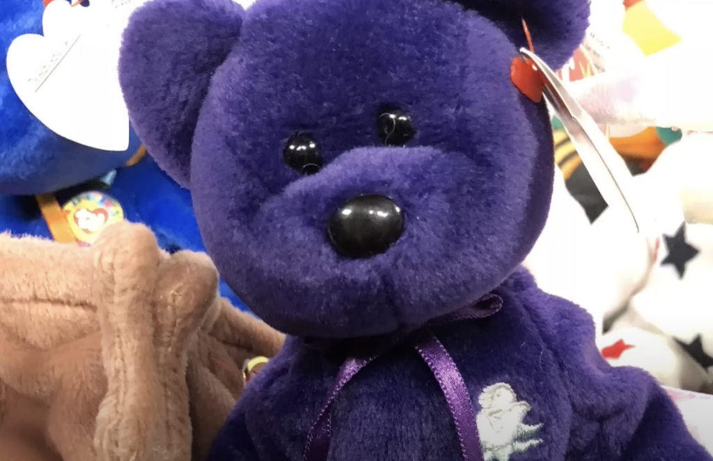 A close-up of a plush, purple teddy bear with black eyes and a small smile. The bear has a ribbon around its neck and is surrounded by other stuffed animals, including a brown one. A heart-shaped tag is attached to its ear.