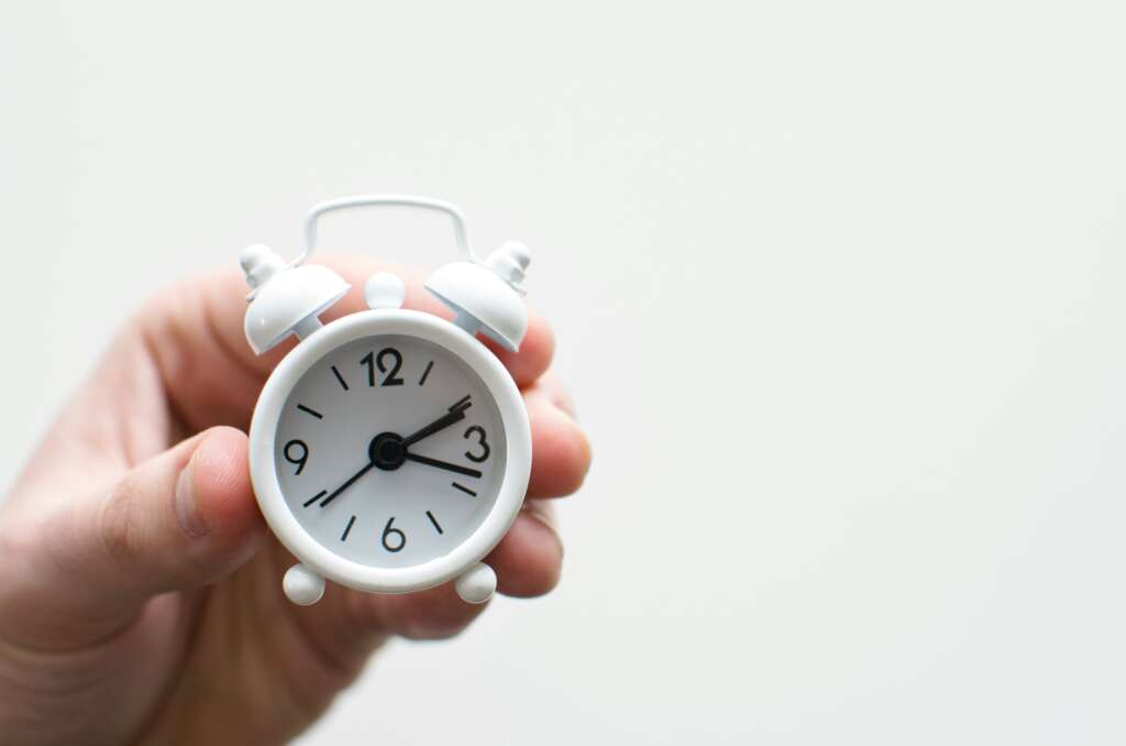 A hand holding a small, white analog alarm clock against a plain background. The clock shows the time as 3:55.